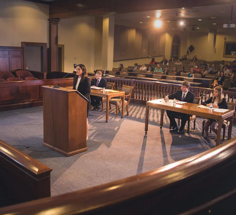 A woman stands at a courtroom podium presenting a case, while suited peers listen attentively and take notes. The setting includes a judge's bench, jury box, and an audience, simulating a real trial environment.