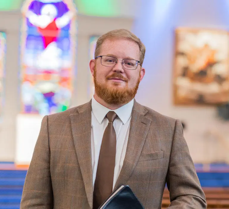 A well-dressed man stands inside a church, holding a Bible and smiling confidently. The background features stained glass windows and soft lighting.