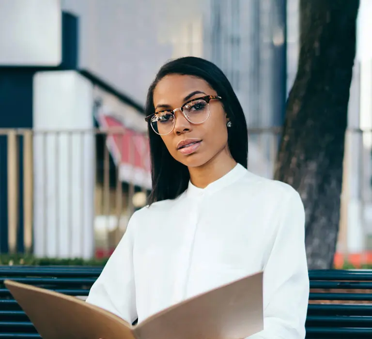 A woman in glasses and a white blouse reads a folder while seated on a bench outdoors.