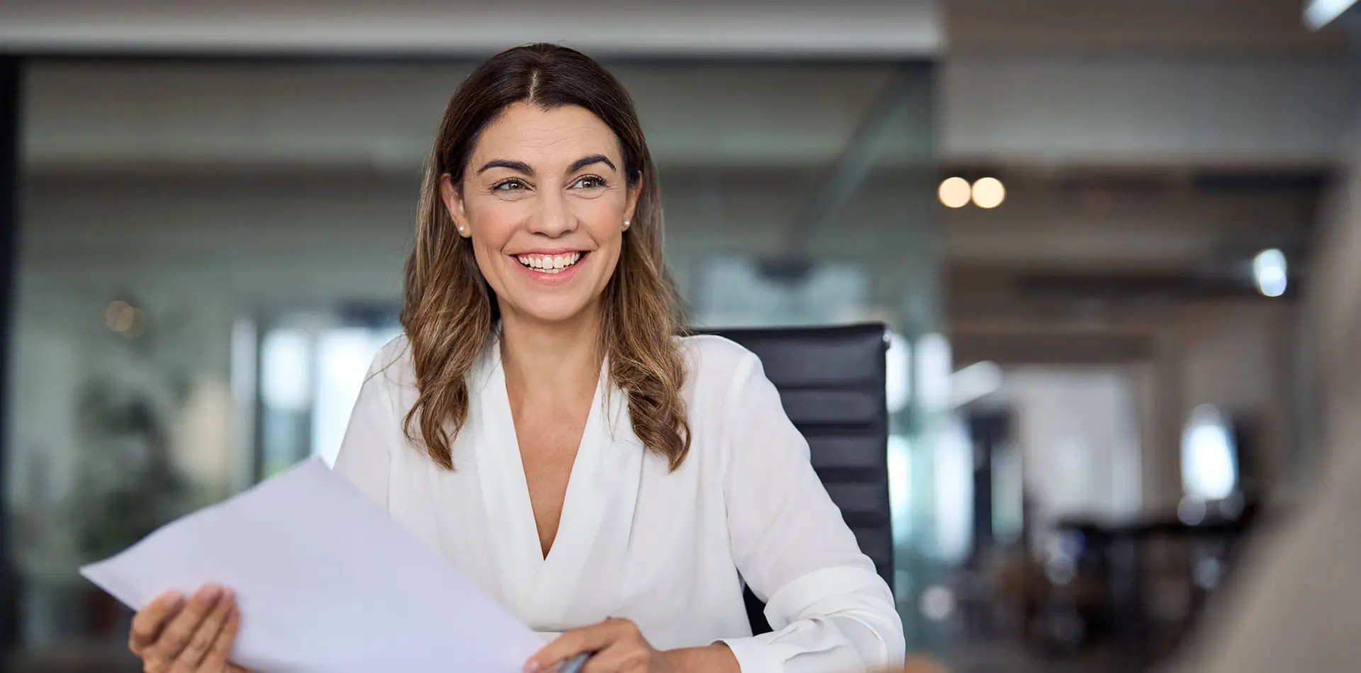 ChatGPT said: A smiling woman in a white blouse holds papers while sitting in a modern office setting.
