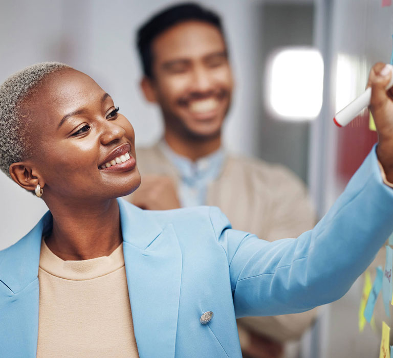 A smiling woman writes on a glass wall covered with sticky notes, with a colleague nearby.